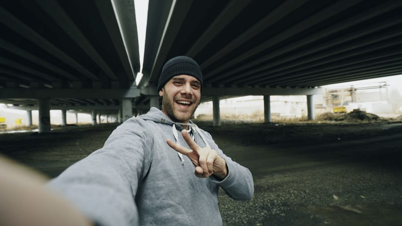 A man in a beanie grinning and throwing a peace sign at his phone camera under a bridge