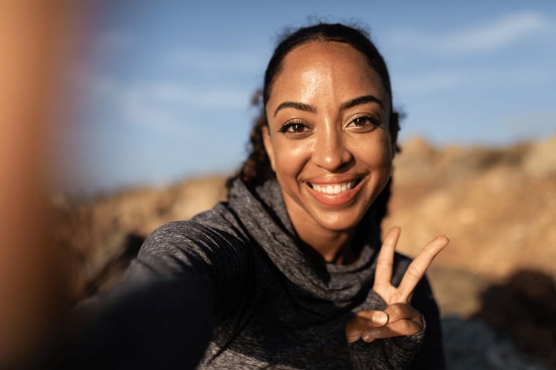 A woman smiling and throwing a peace sign at her phone camera outdoors