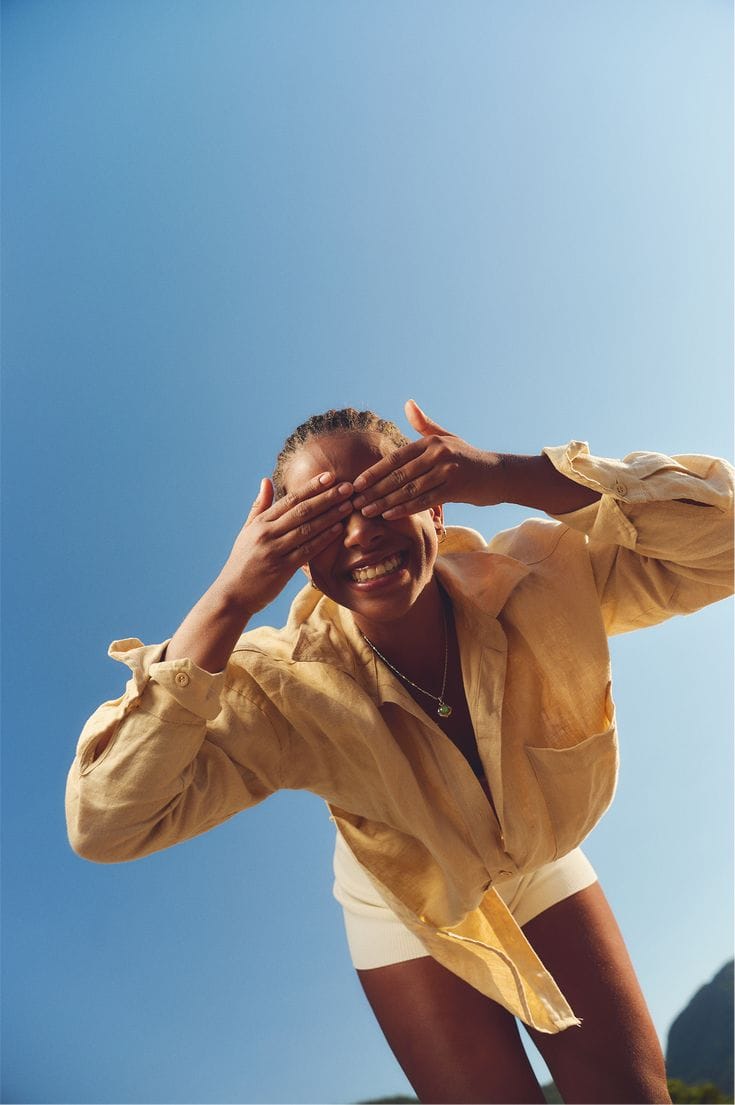 A woman in a yellow jacket shading her eyes and laughing against a clear sky
